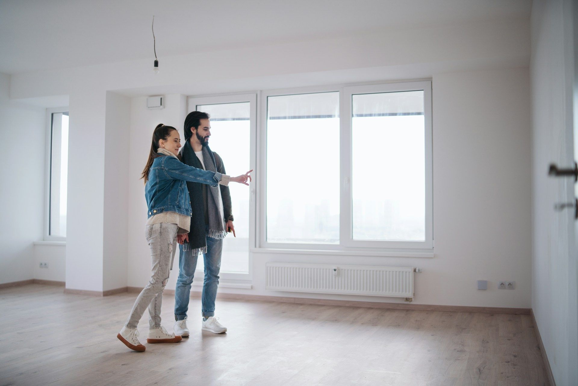 Couple inspecting a bright, empty room with large windows, gesturing and looking around.