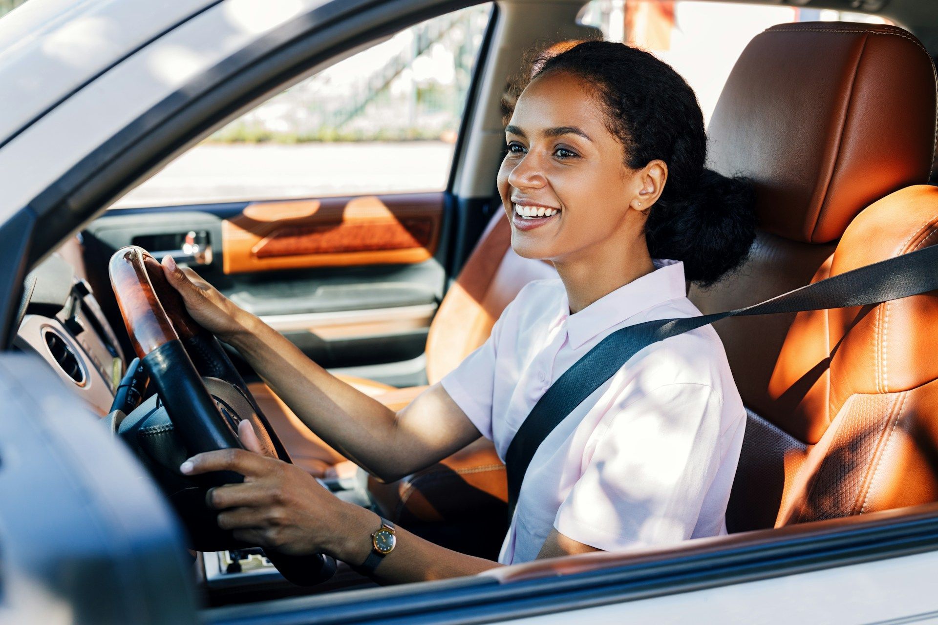 Woman smiling while driving a car, wearing seatbelt, sunlight, leather interior.