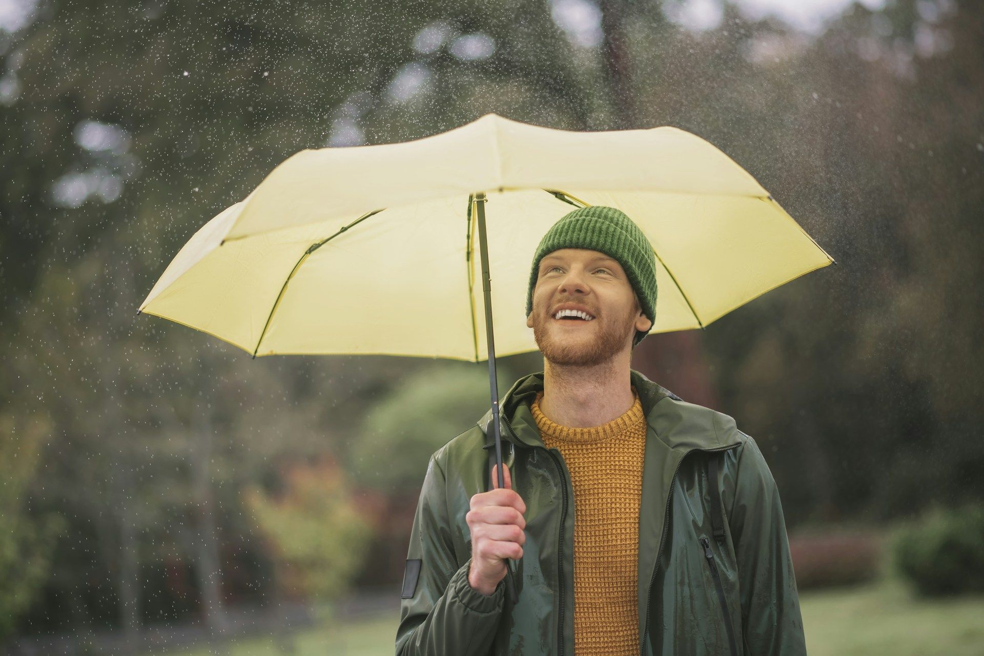 Man in green jacket and hat smiles while looking up at rain under a yellow umbrella.