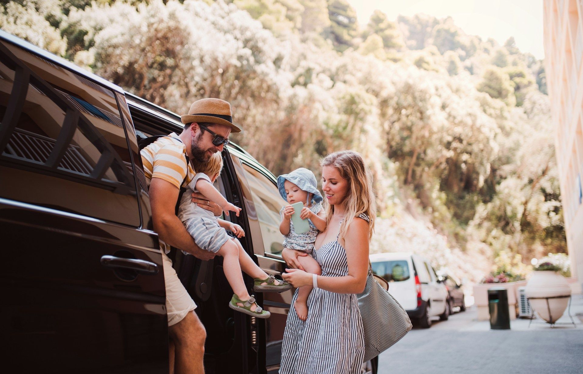 Family exits a black vehicle on a street lined with trees and parked cars.