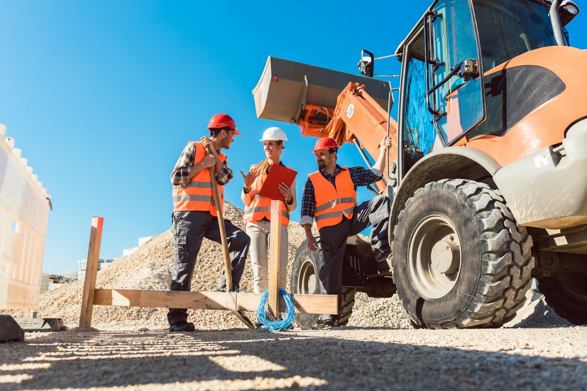 Construction workers in orange vests and hard hats, near machinery and a pile of stones.