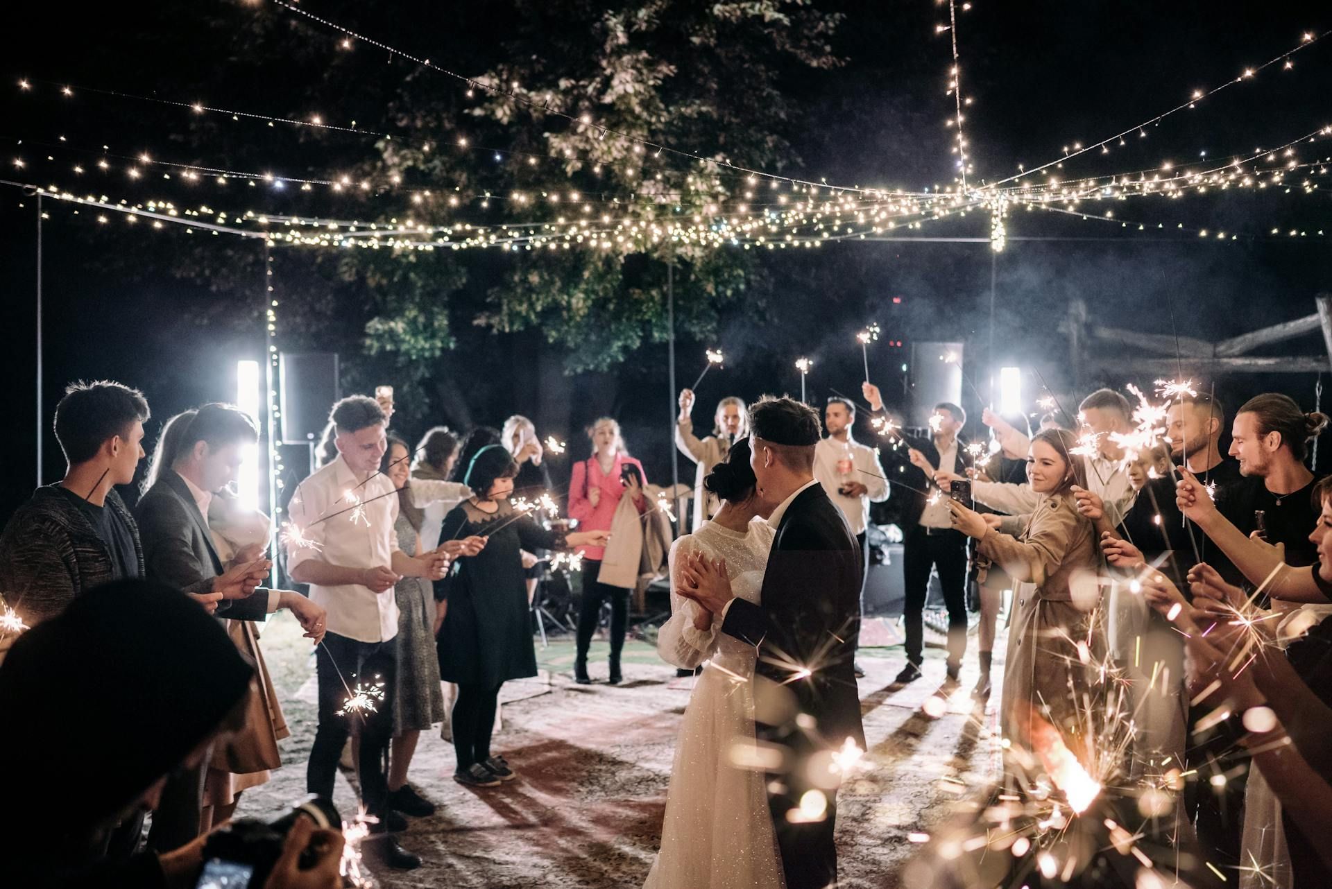 Couple dances surrounded by guests holding sparklers under string lights.