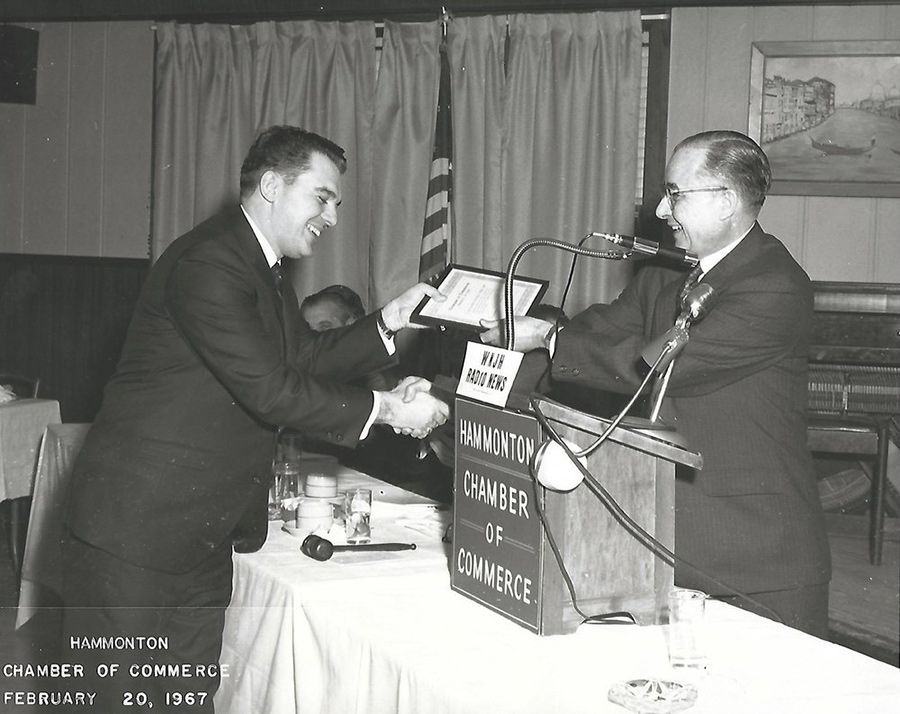 Two men shaking hands in front of a chamber of commerce sign