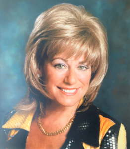 Woman with blonde hair, smiling, wearing a gold chain and sequined jacket.