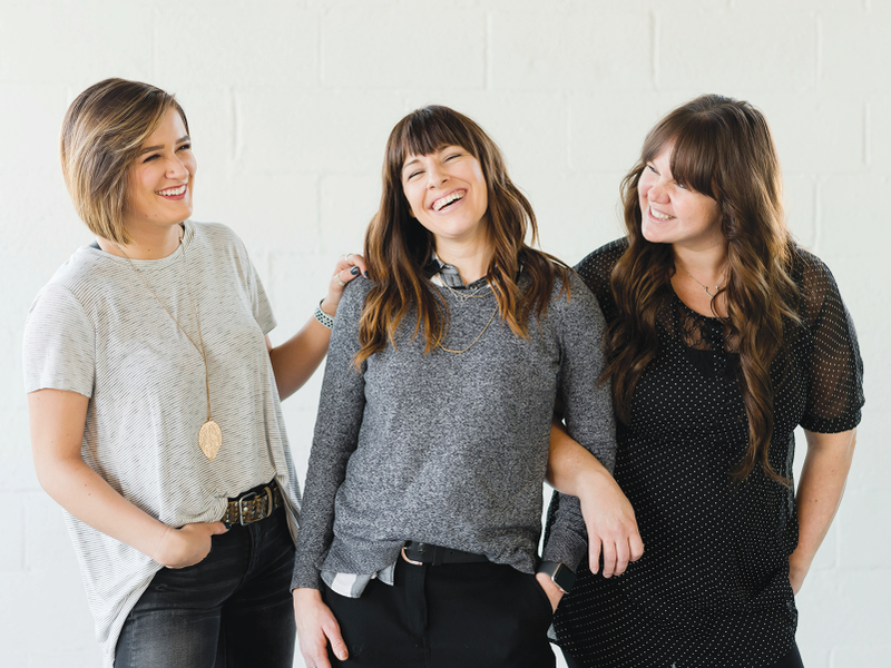 Three women smiling together in front of a white wall. One has her arm around another's shoulders.