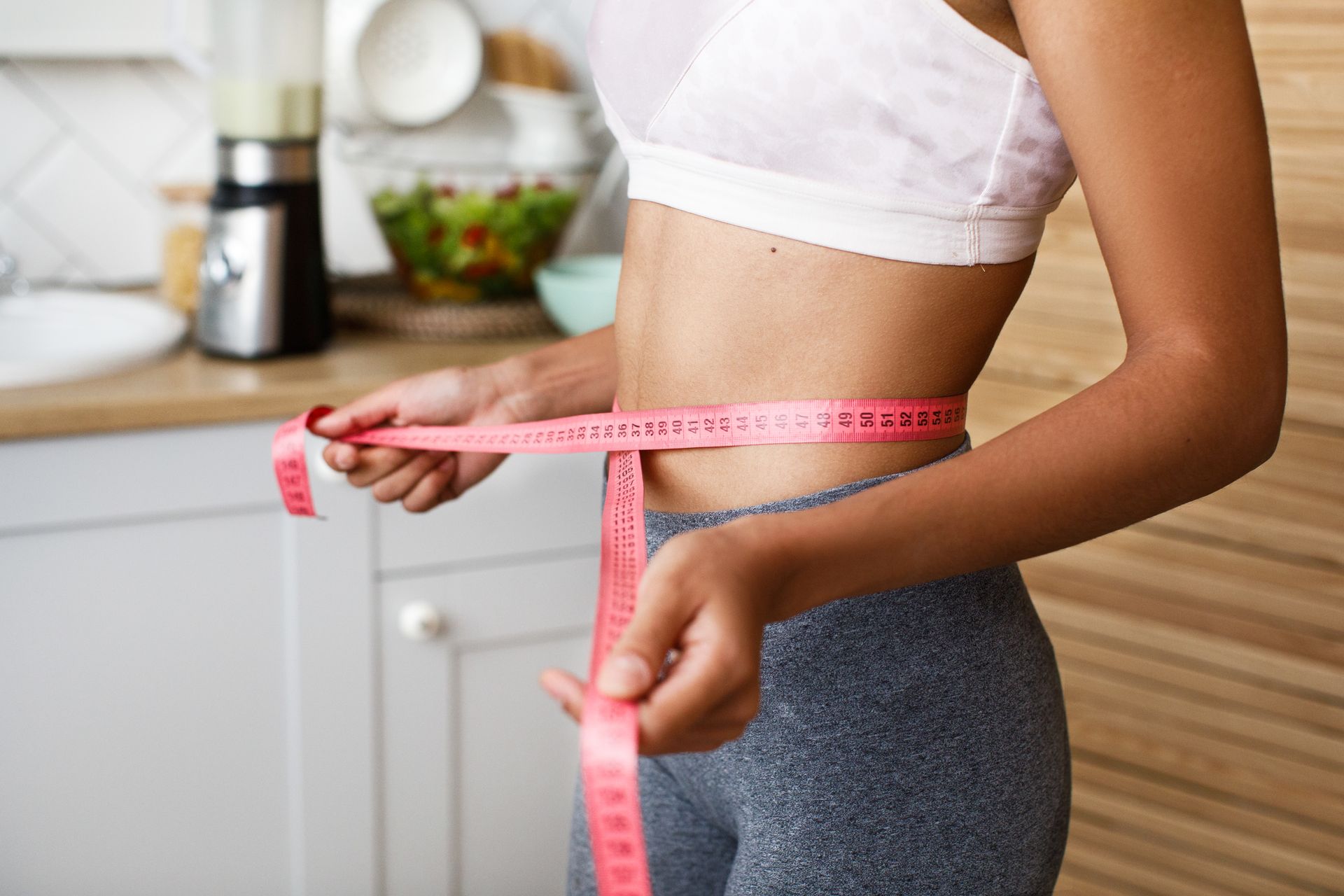 A woman is measuring her waist with a tape measure in a kitchen.