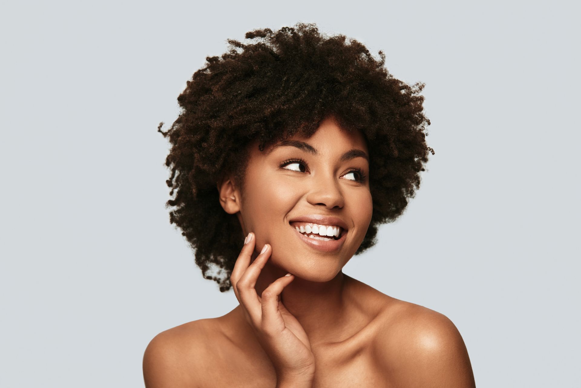 A woman with curly hair is smiling and touching her face.