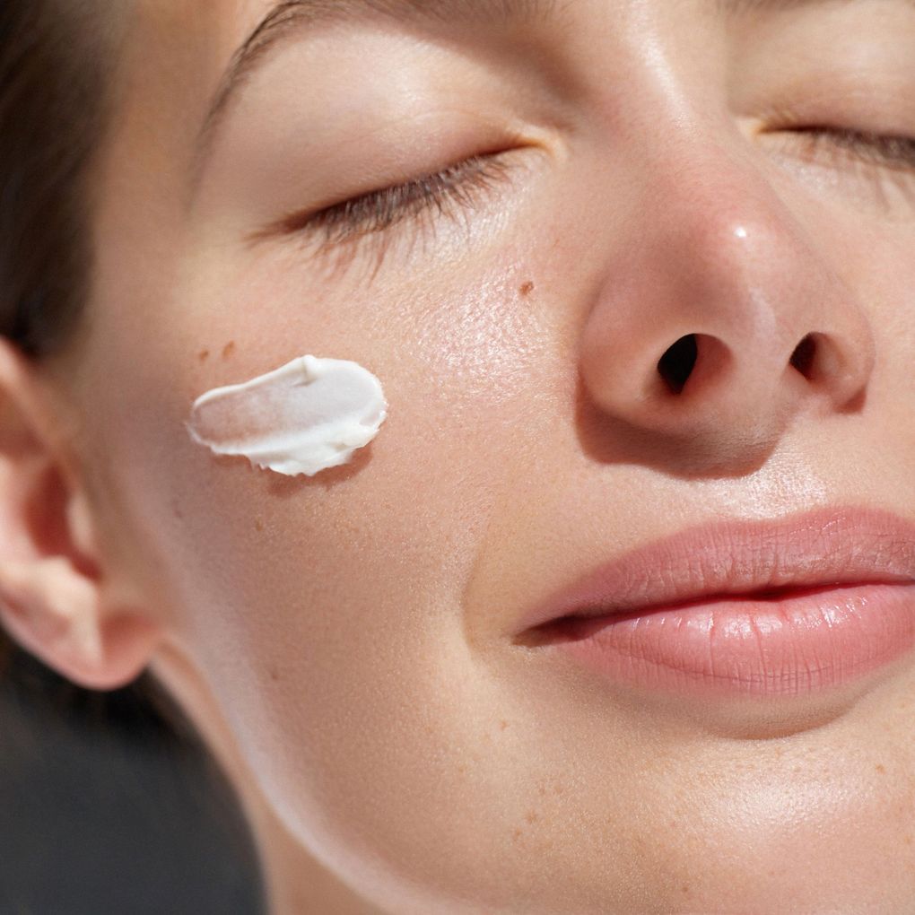 A woman is applying cream to her face with her eyes closed.