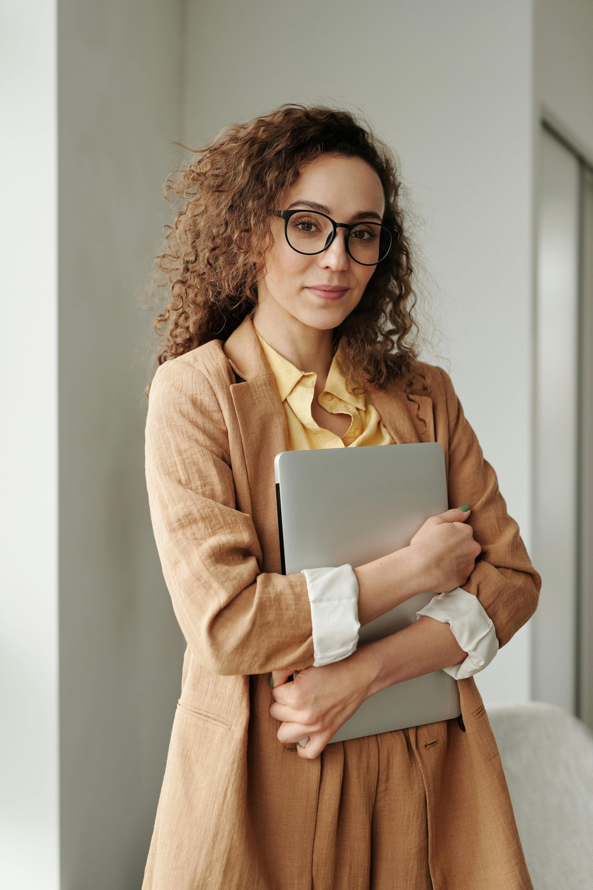 Woman with curly hair and glasses, holding laptop, wearing blazer and yellow shirt, indoors.