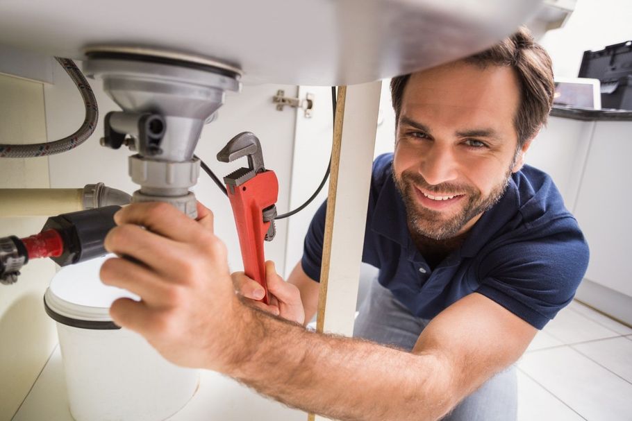 Man repairing a sink drain with a wrench under a bathroom counter, smiling at the camera