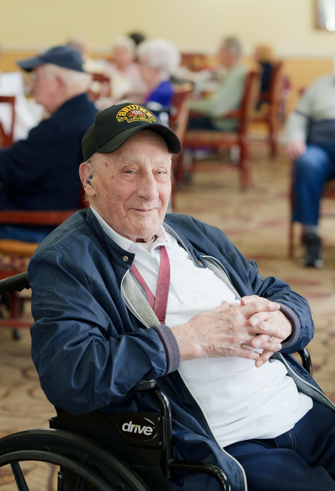 An individual in a wheelchair and a baseball cap smiles in a room with other people seated at tables in the background.