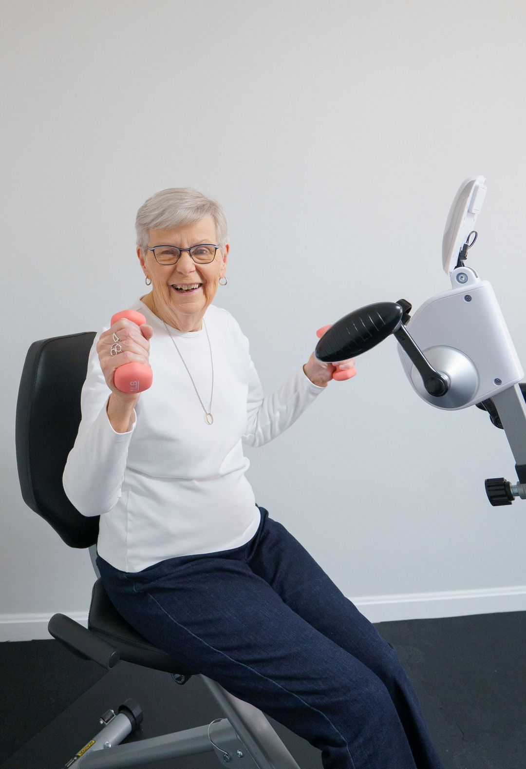 An elderly woman is sitting on an exercise bike holding dumbbells.