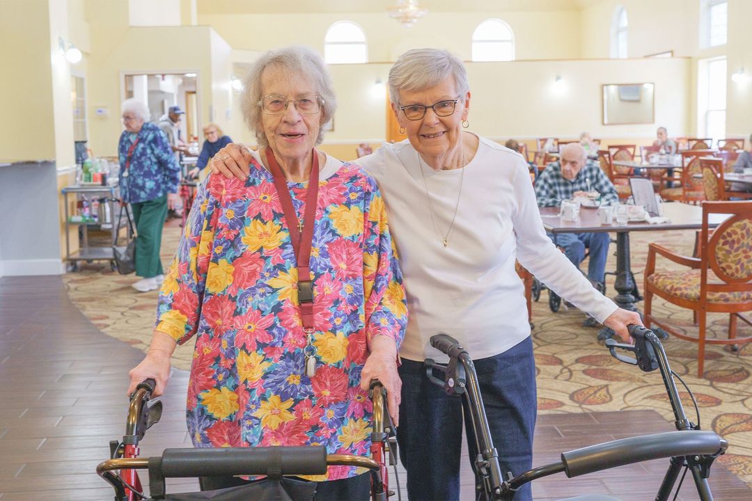 Two elderly women are posing for a picture in a nursing home . one of the women is using a walker.