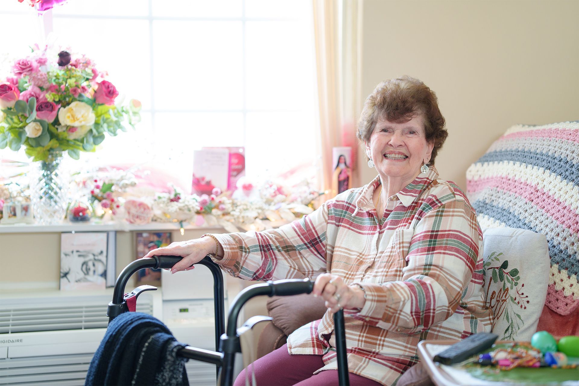 An elderly woman is sitting in a chair with a walker and smiling.