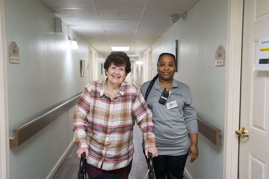 A woman with a walker is walking down a hallway next to a nurse.