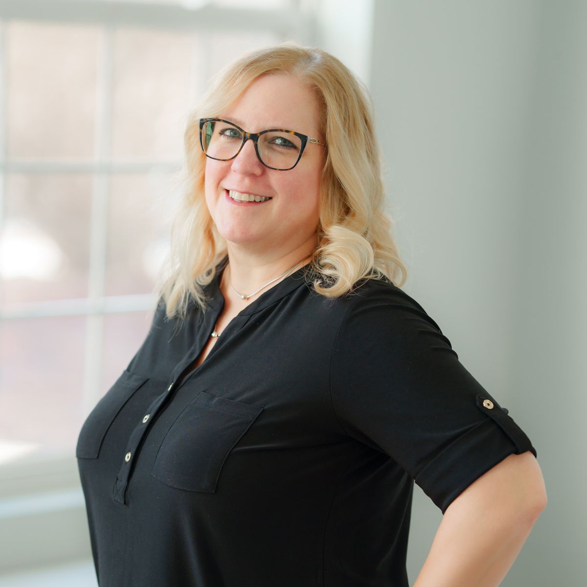 A woman wearing glasses and a black shirt is standing in front of a window.