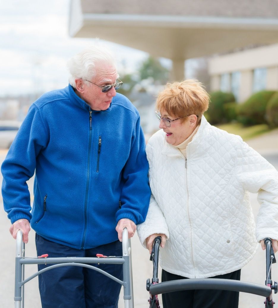 Elderly couple using walkers, outdoors. Man in blue jacket, woman in white jacket. They are looking at each other, possibly talking.
