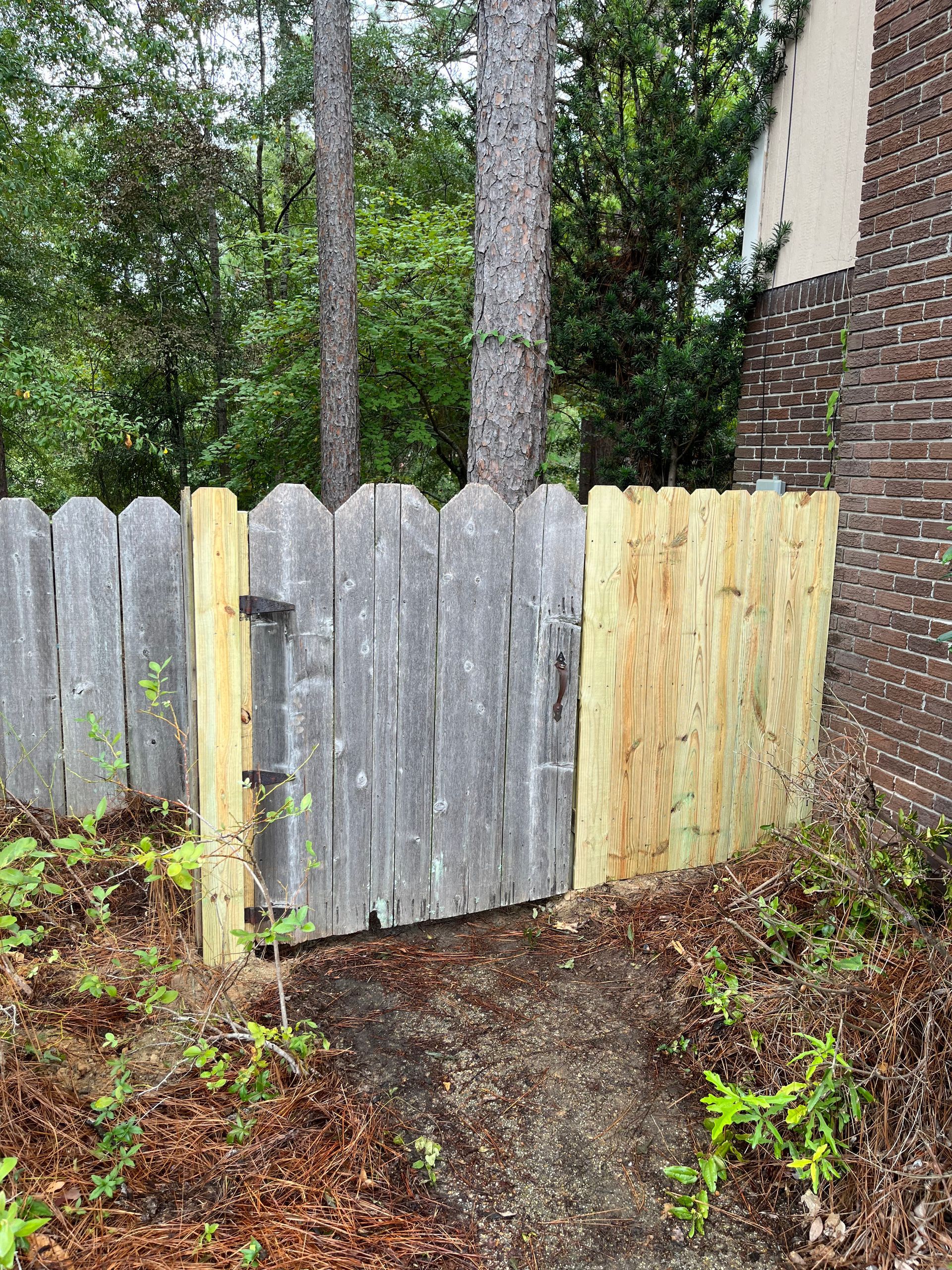 A wooden fence with a wooden gate is surrounded by trees and a brick building.