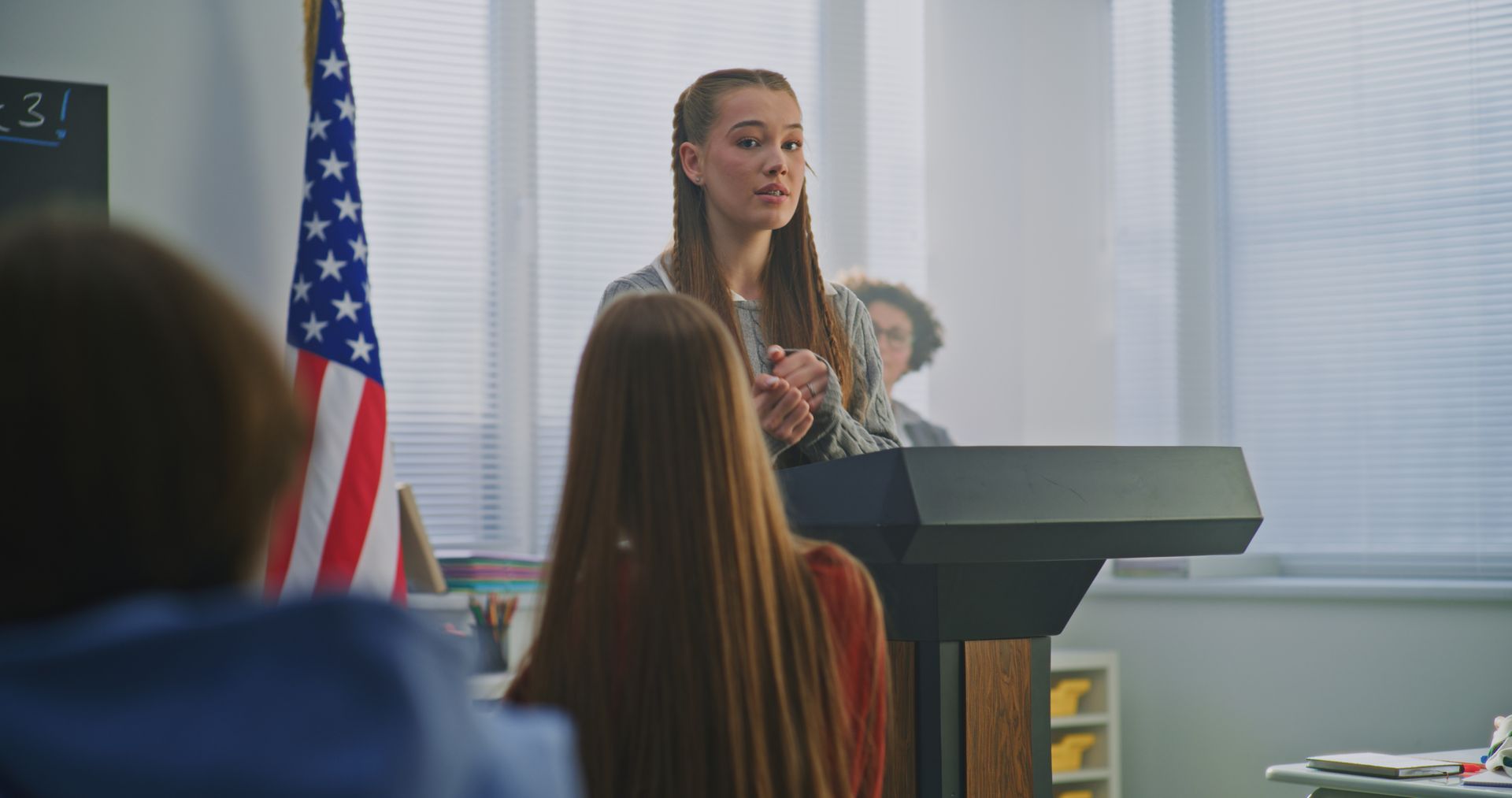 Speaker at a podium addressing an audience in a meeting room, with an American flag nearby.