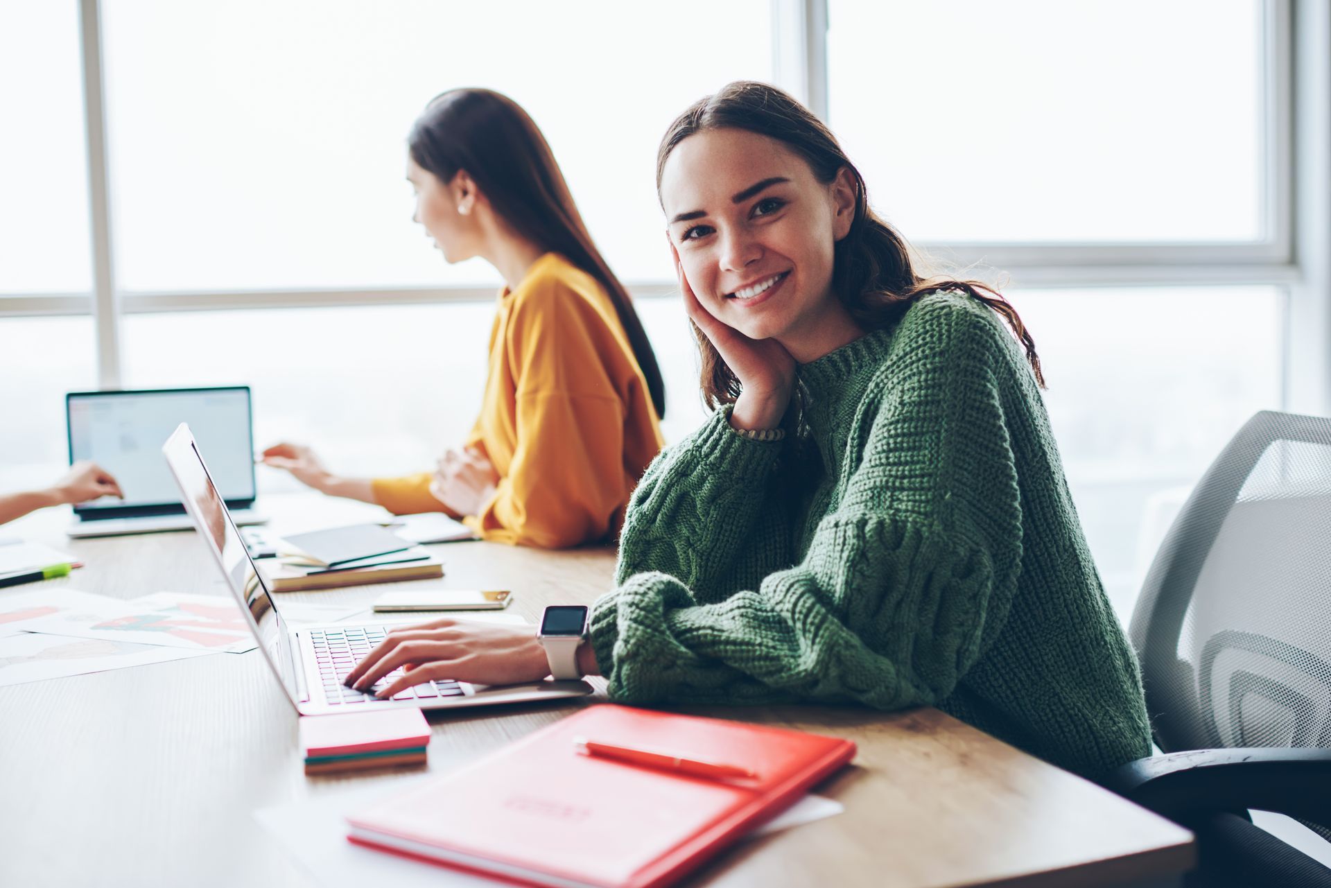 A smiling person in a green sweater uses a laptop at an office desk, while another person works in the background.