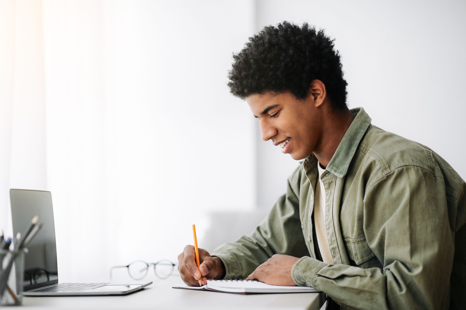 Person writing in a notebook at a desk, smiling near a laptop and glasses.