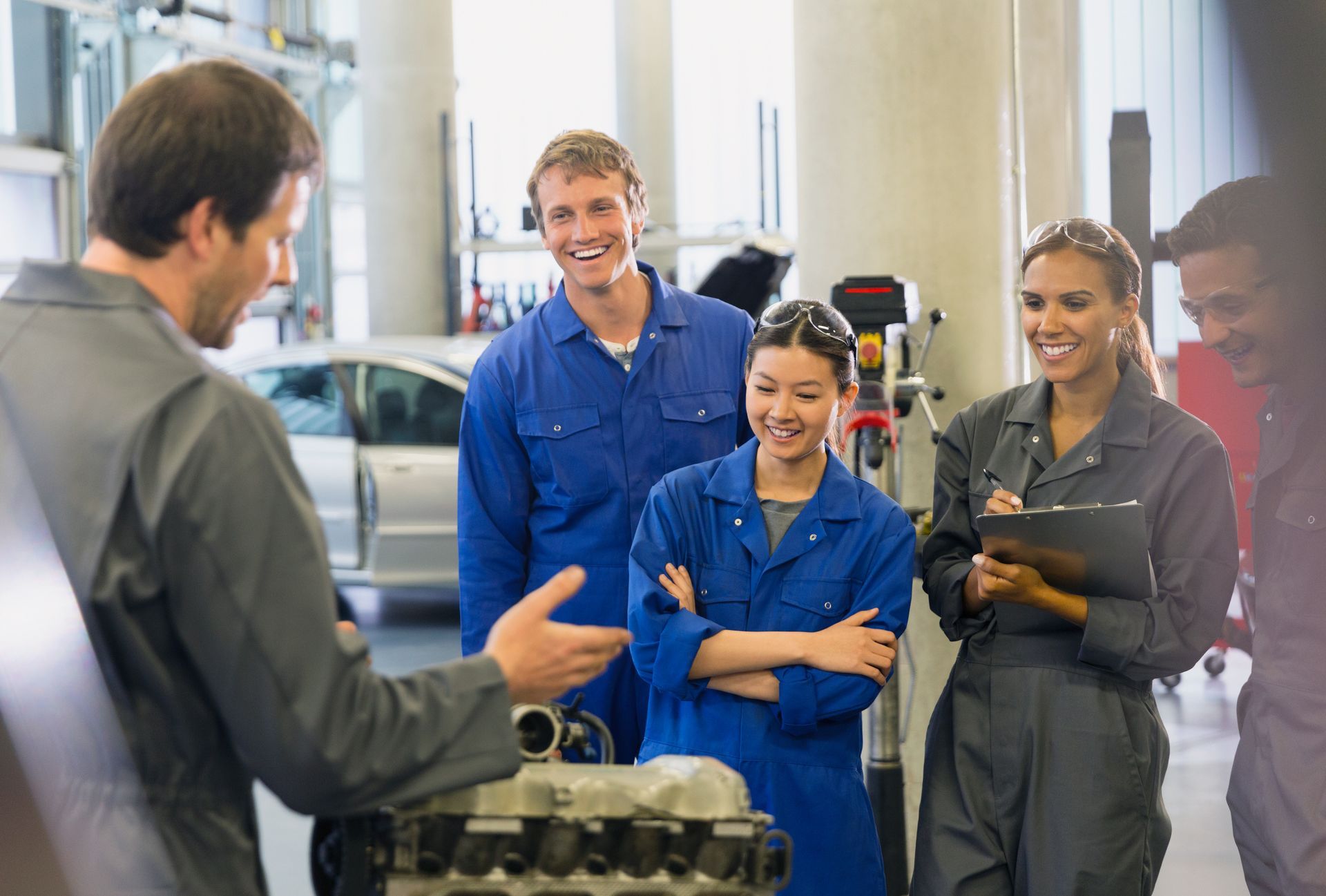 A group of mechanic students are standing around a car engine in a garage.