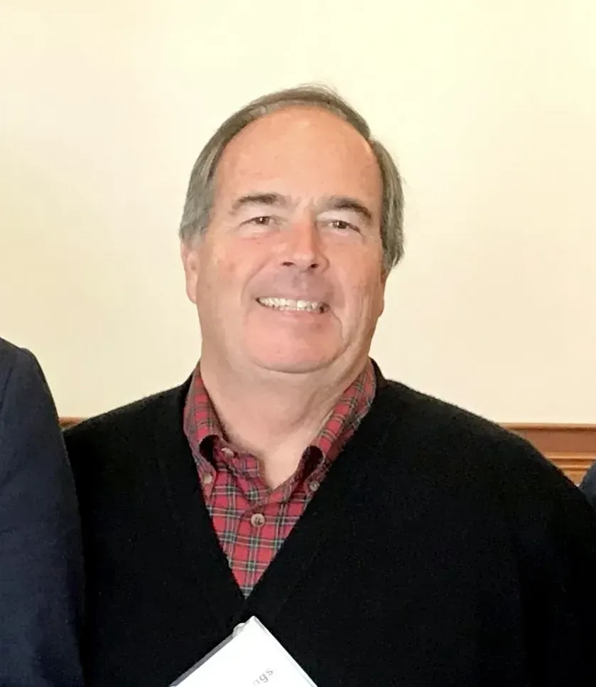 A man wearing a black sweater and a plaid shirt is smiling for the camera.