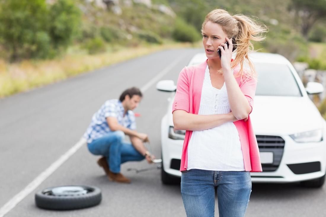 A man is changing a tire on a car while a woman talks on a cell phone.