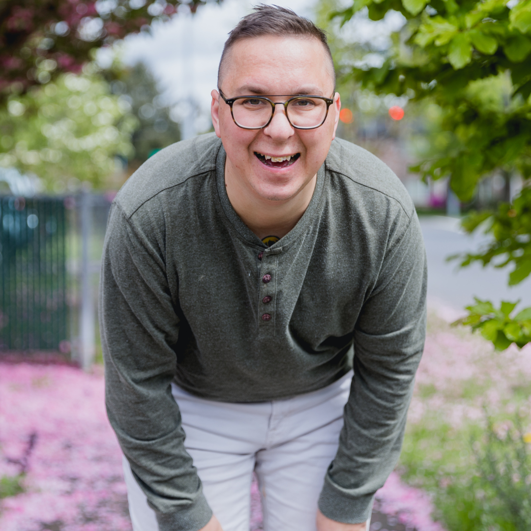 Man wearing glasses, olive green shirt, and white pants, smiling outside near pink flowers.