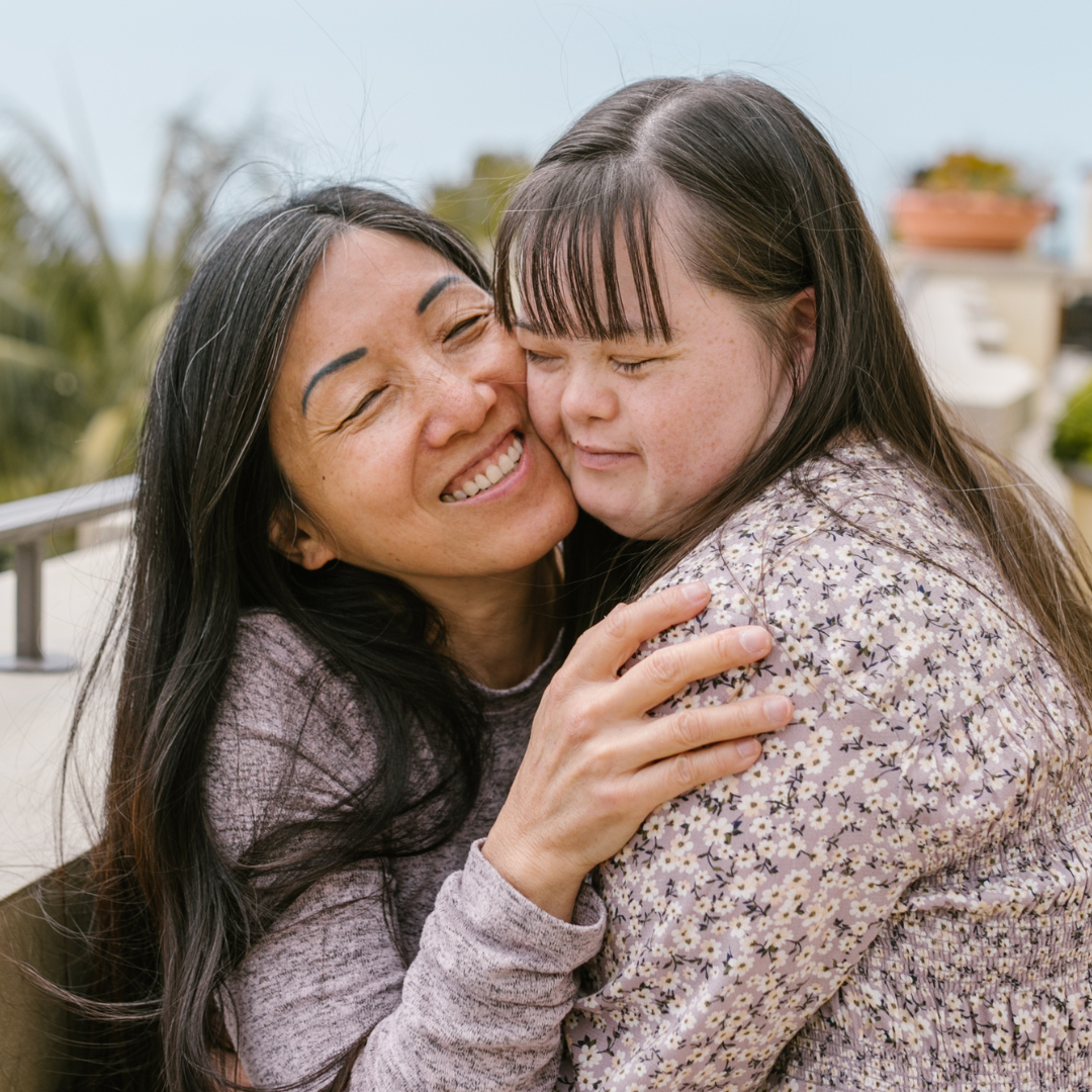 Asian woman hugs a person with Down syndrome, both smiling. Outdoors on a balcony, neutral tones.