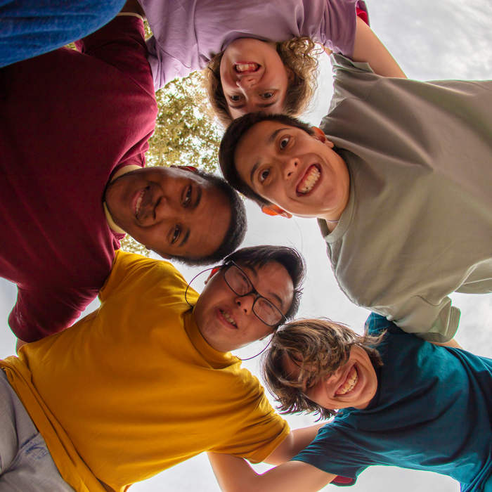Group of people smiling, heads together in a circle, viewed from below; outdoors.