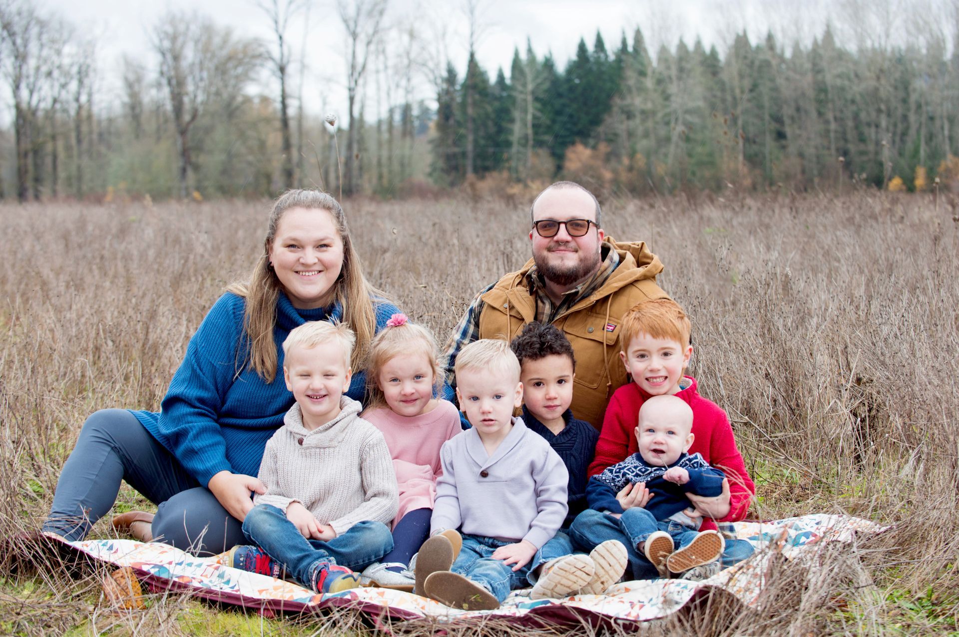 Family of nine poses on a blanket in a field: a woman, a man, and seven children. They smile in front of a forest.