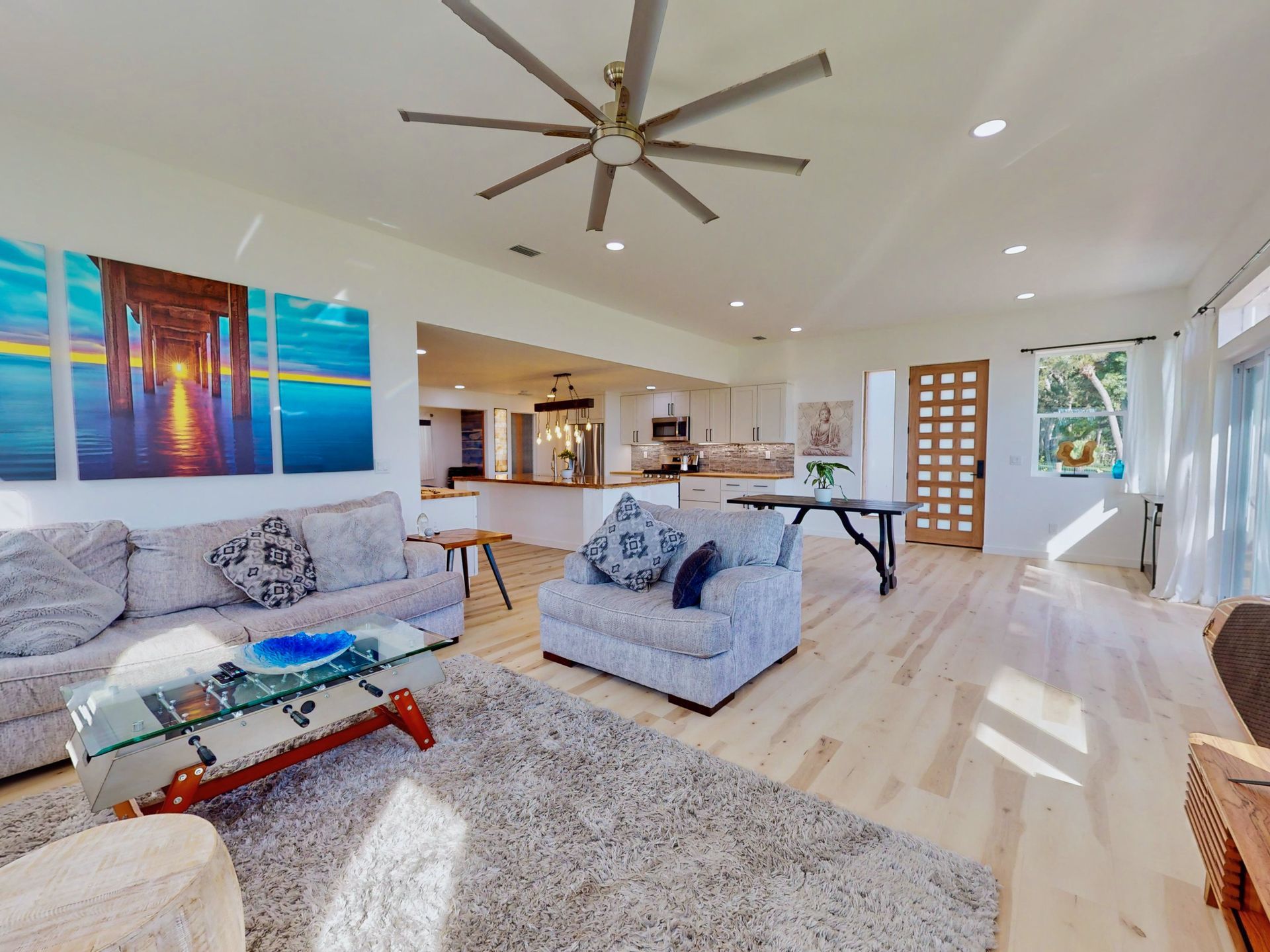 A bright living room with light wood floors, a sofa, patterned armchair, a glass coffee table, and a large ocean wall art.