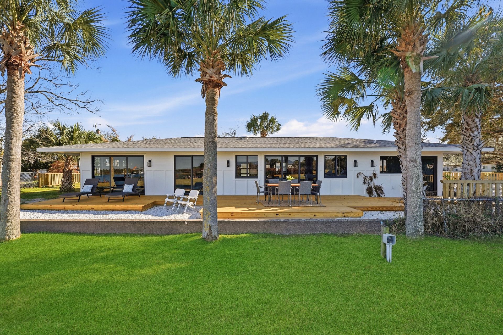 A white, single-story house with a wooden deck and large sliding glass doors, framed by palm trees and a green lawn.