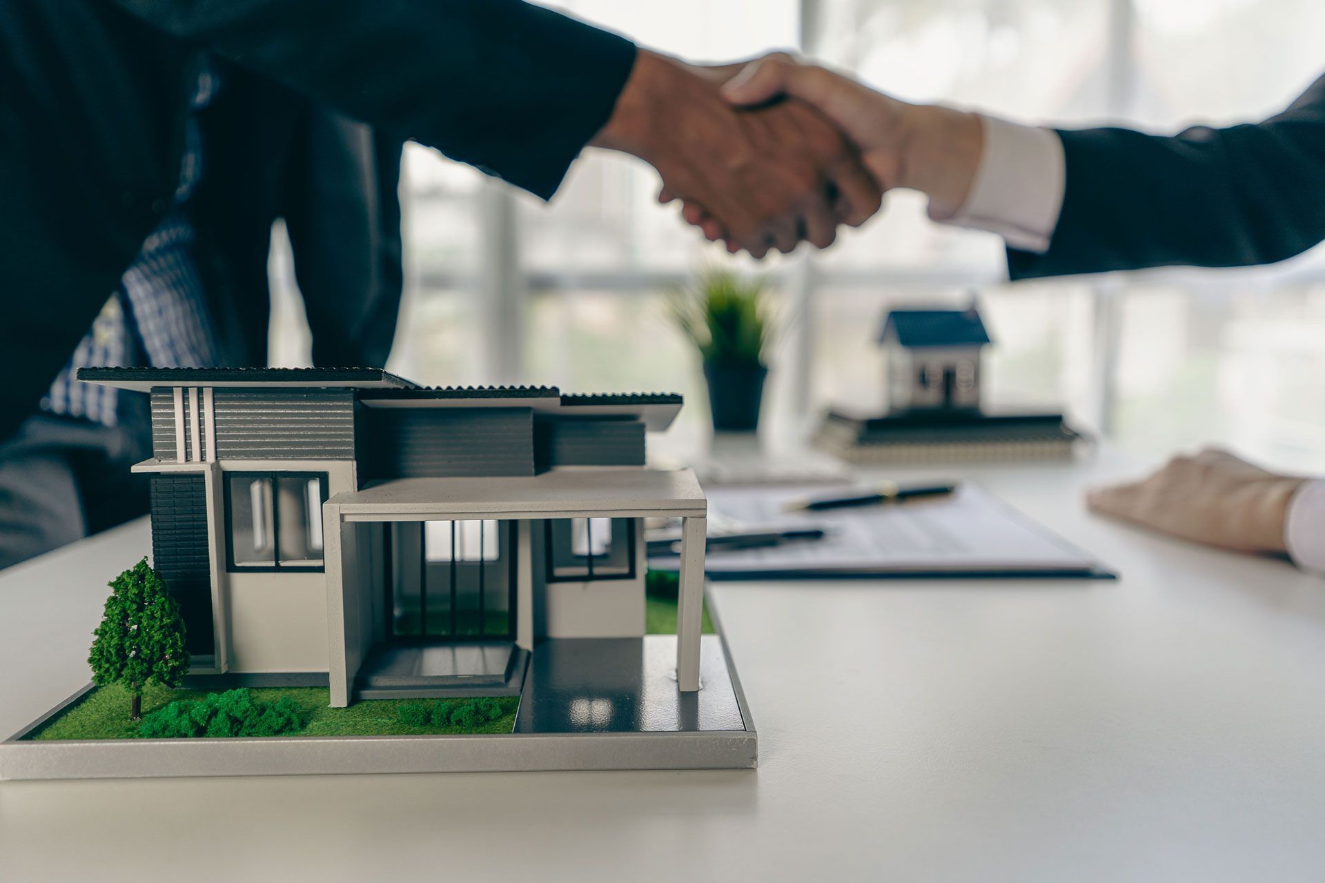 Two people shake hands over a table with a model house, representing a real estate deal or home purchase.