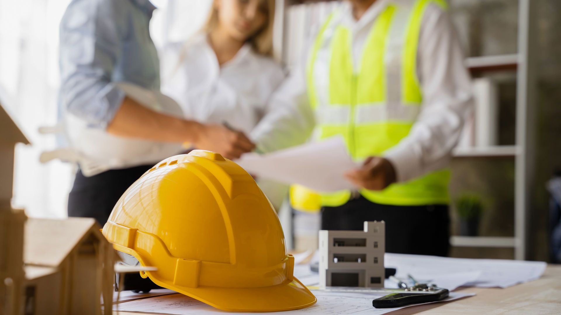A yellow hard hat rests on a table with architectural models, as professionals review blueprints in the background.
