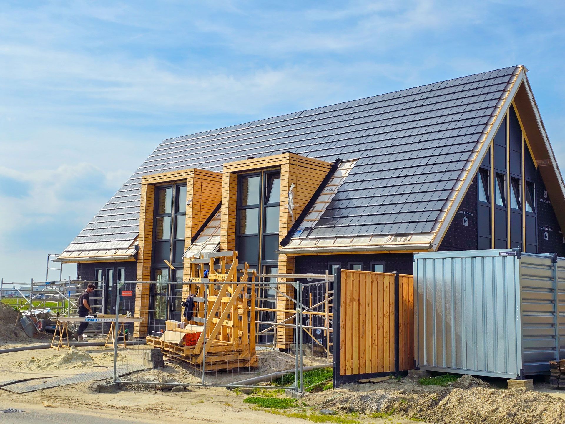 A house under construction with a dark tiled roof, wooden framing around tall windows, and a metal storage container nearby.