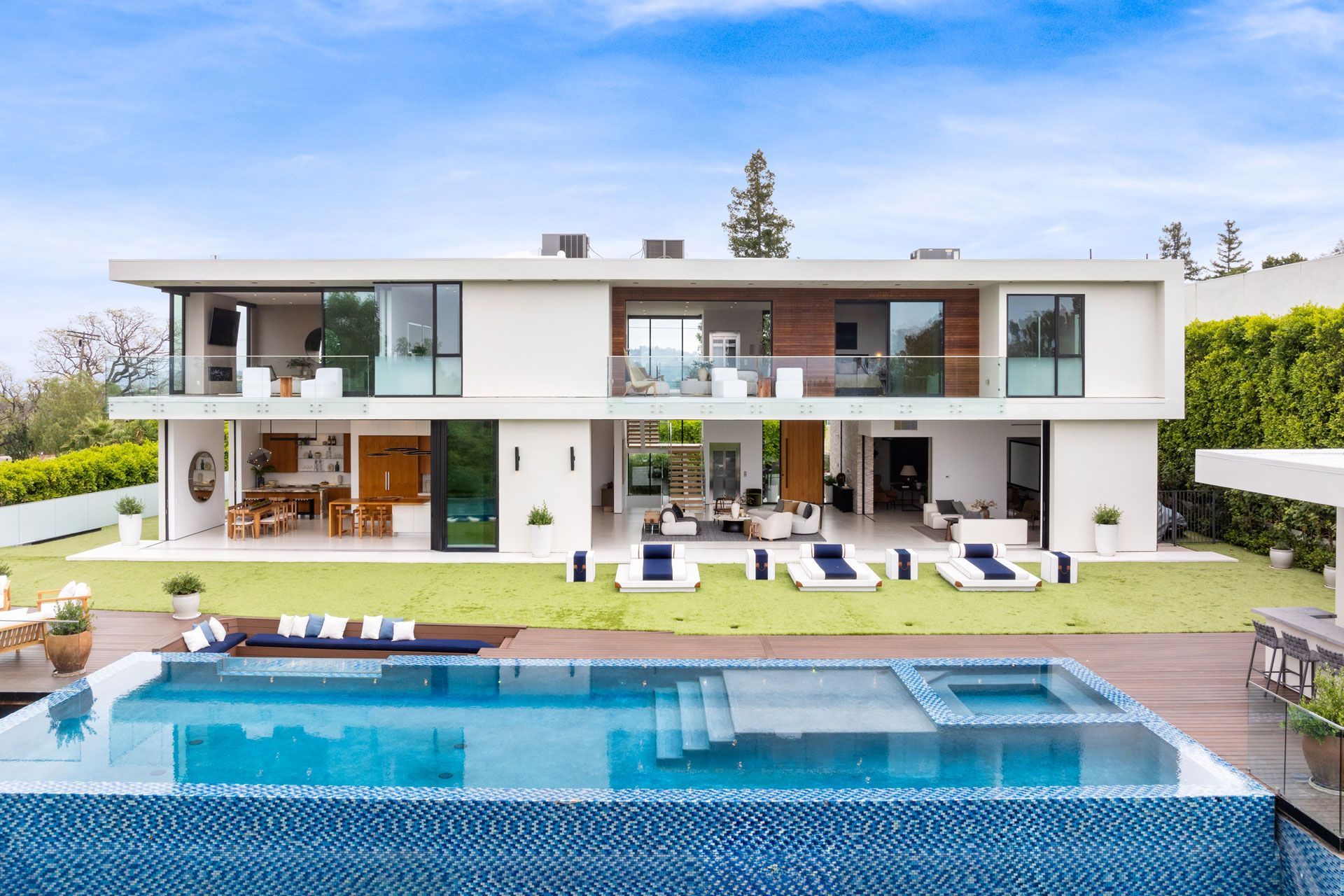A modern two-story home with a white exterior, large sliding glass doors, a flat roof, and an infinity pool in the yard.
