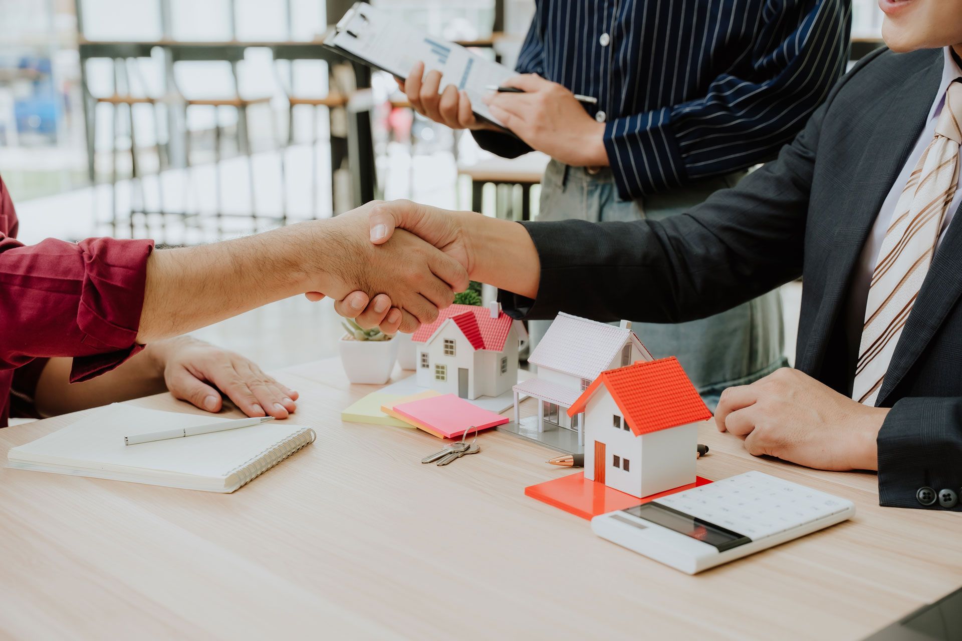 Two professionals shake hands over a table featuring miniature house models and financial documents in an office setting.