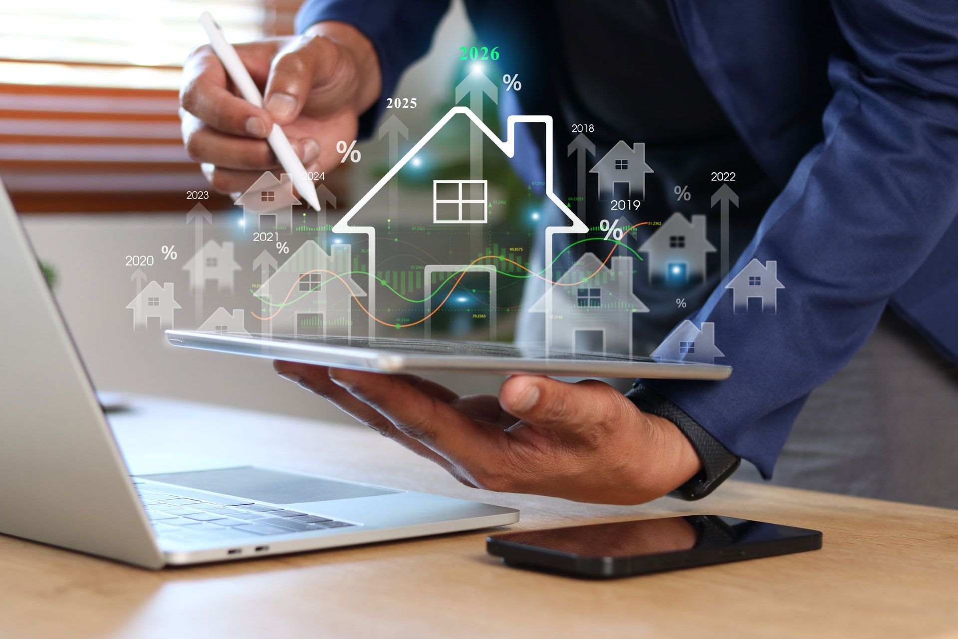 A person using a stylus on a tablet displaying augmented reality icons of houses, with a laptop and phone on the desk.