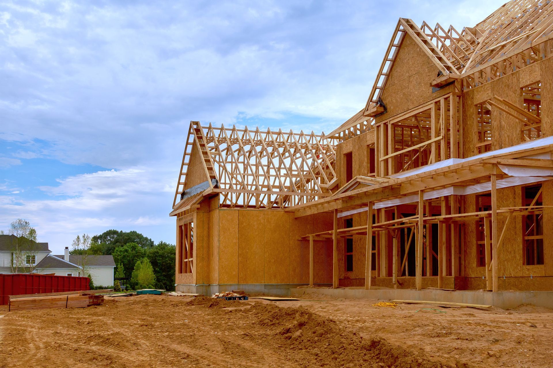 A wooden frame structure of a large house under construction on a dirt lot against a blue, cloudy sky.