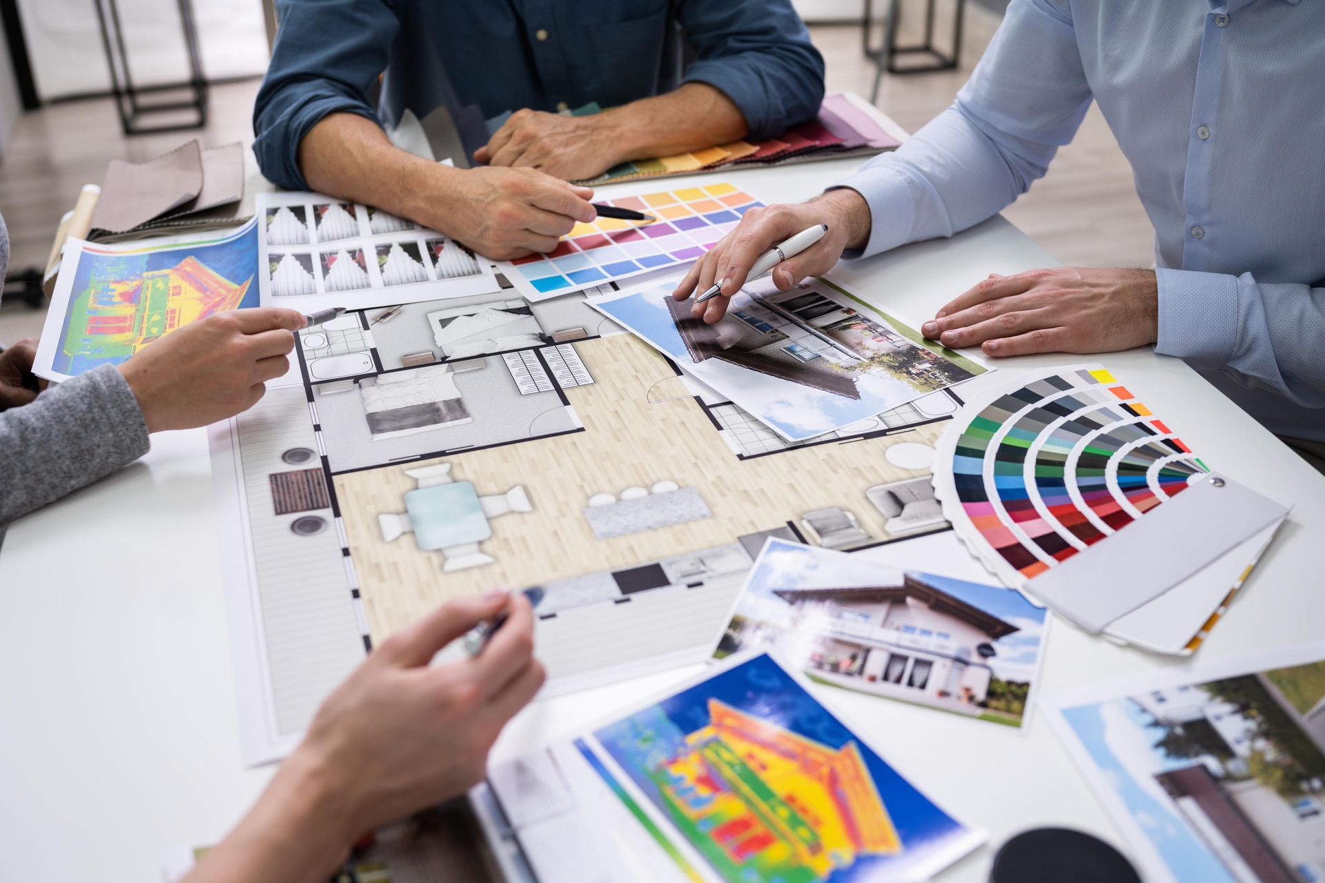 People collaborate around a table, reviewing house floor plans, exterior photographs, and paint swatches for a project.