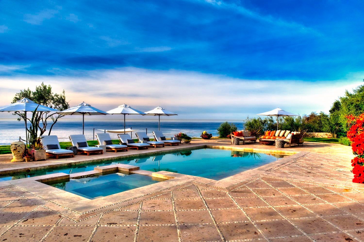A large swimming pool surrounded by chairs and umbrellas with a view of the ocean.