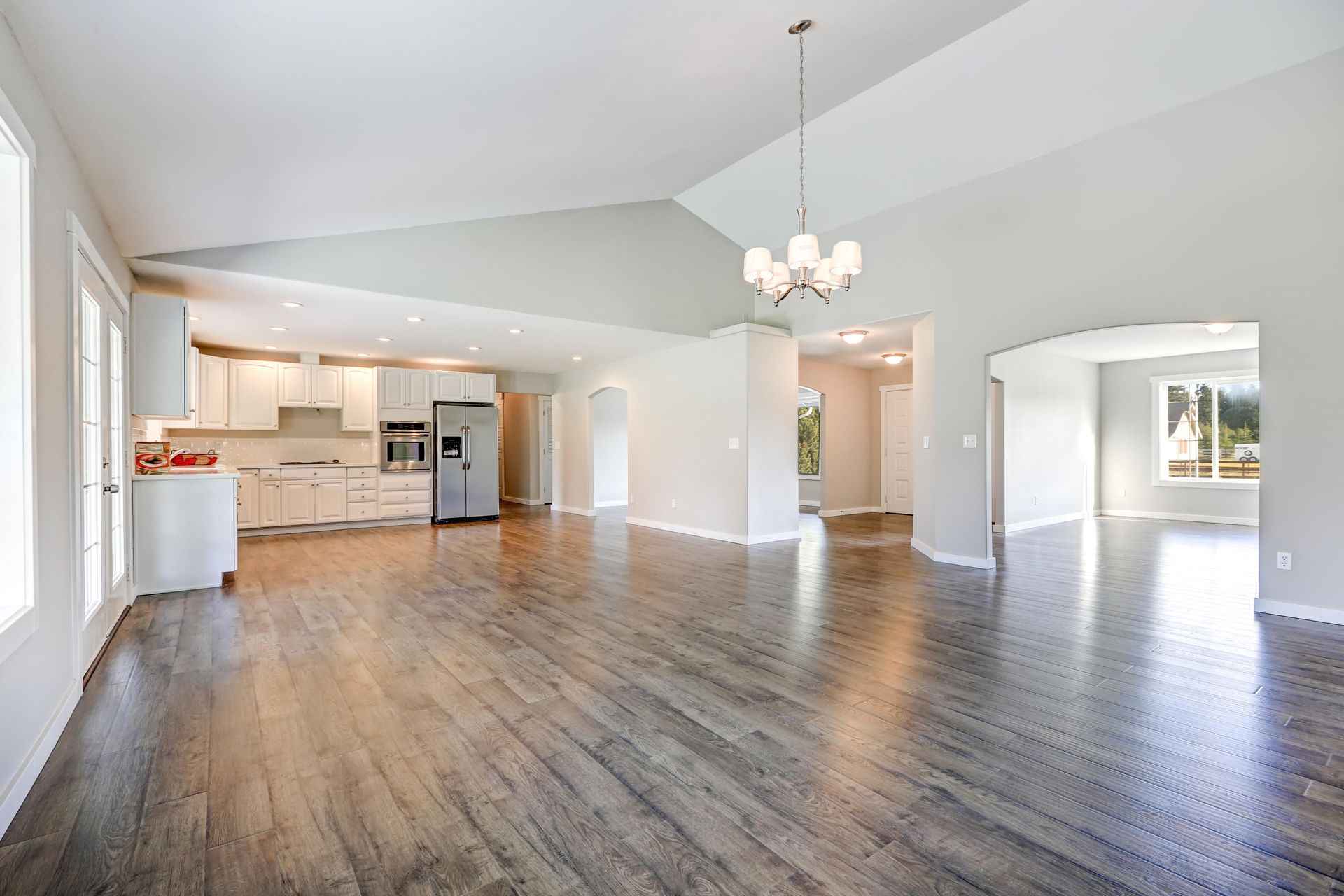 A bright, open-concept living area with a high ceiling, chandelier, light walls, and dark wood-look flooring.