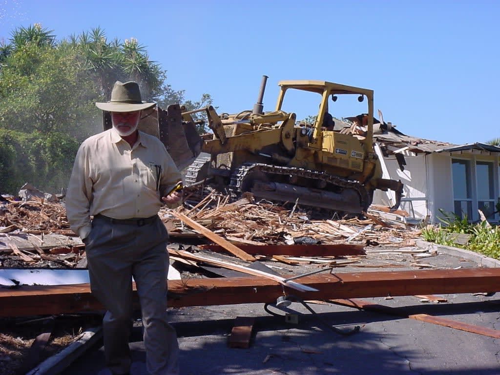 A man in a cowboy hat stands in front of a bulldozer demolishing a house.