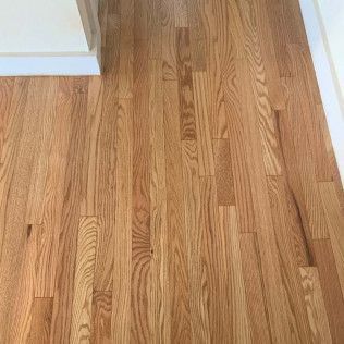 Hardwood floor planks, light brown with wood grain, next to white baseboard and wall.