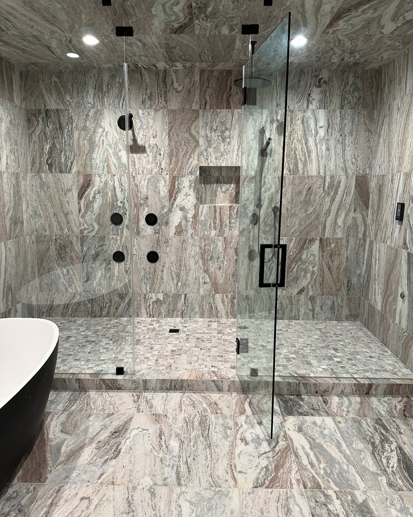 Spacious, marble-tiled shower with glass door and black fixtures.