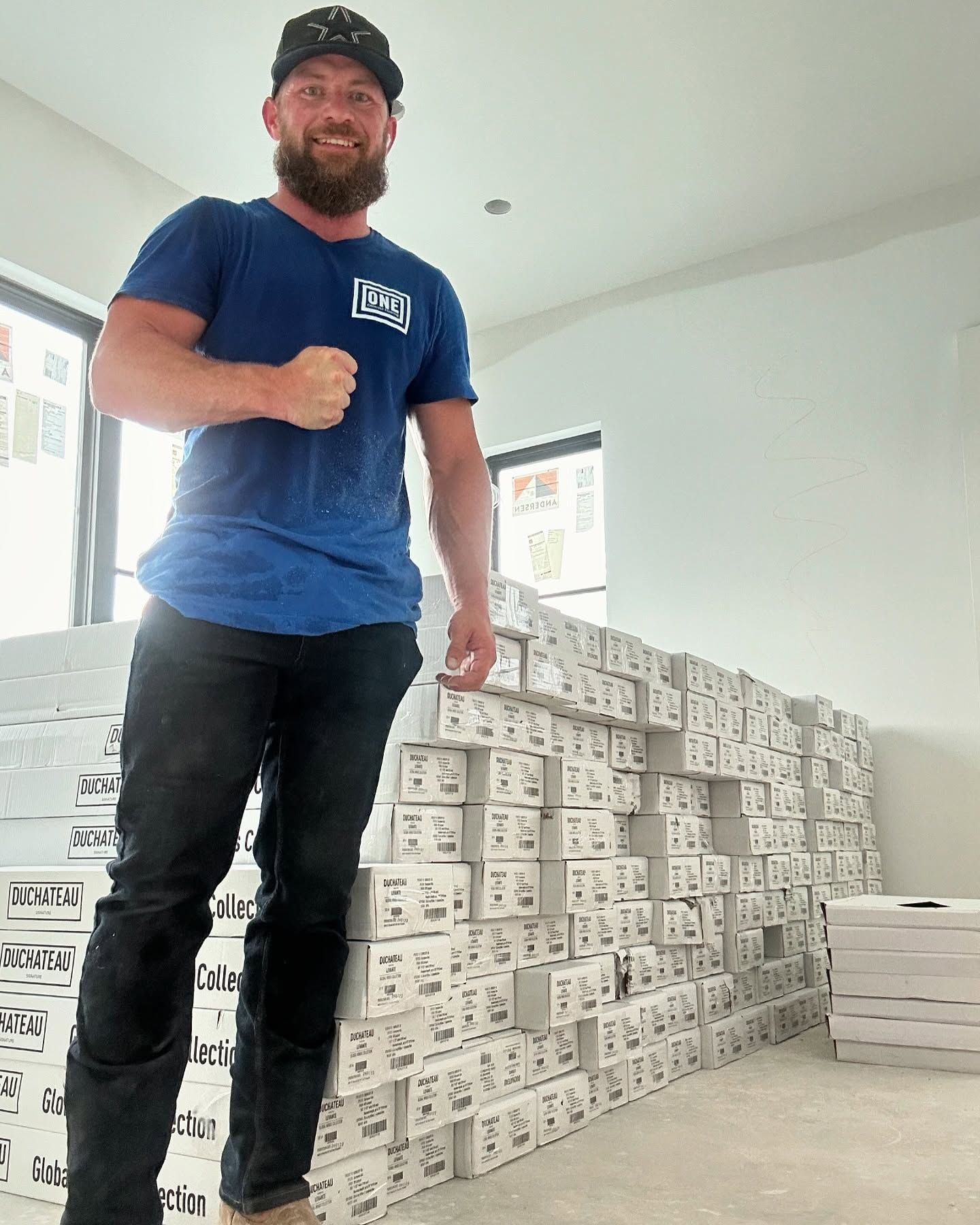 Man in blue shirt, black pants, standing next to a tall stack of boxes in a construction setting.