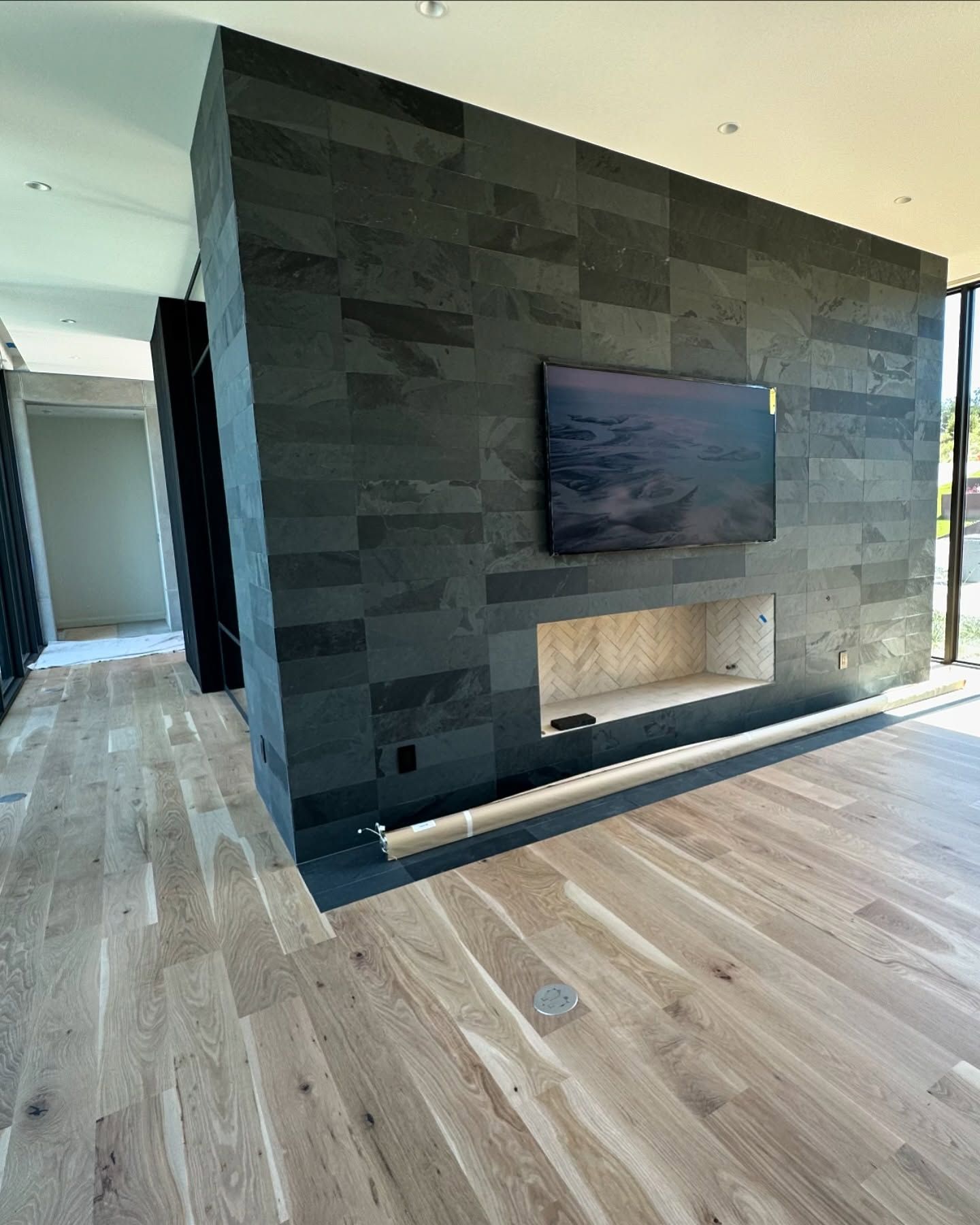 Gray tiled wall with built-in fireplace and TV, oak flooring in a modern room with large windows.