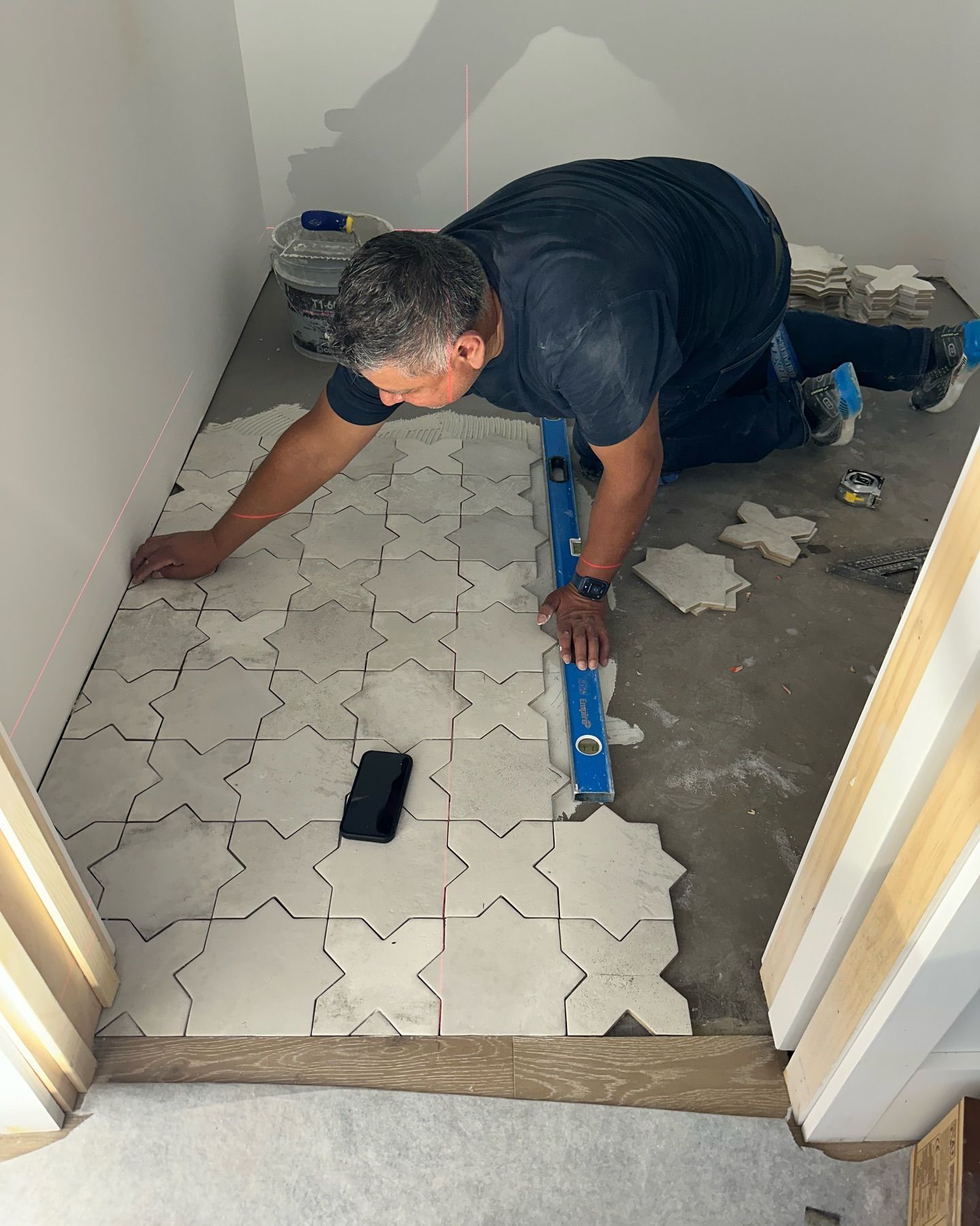Person kneeling, laying tile floor in room. Tiles are light-colored, in a star shape. Level used.